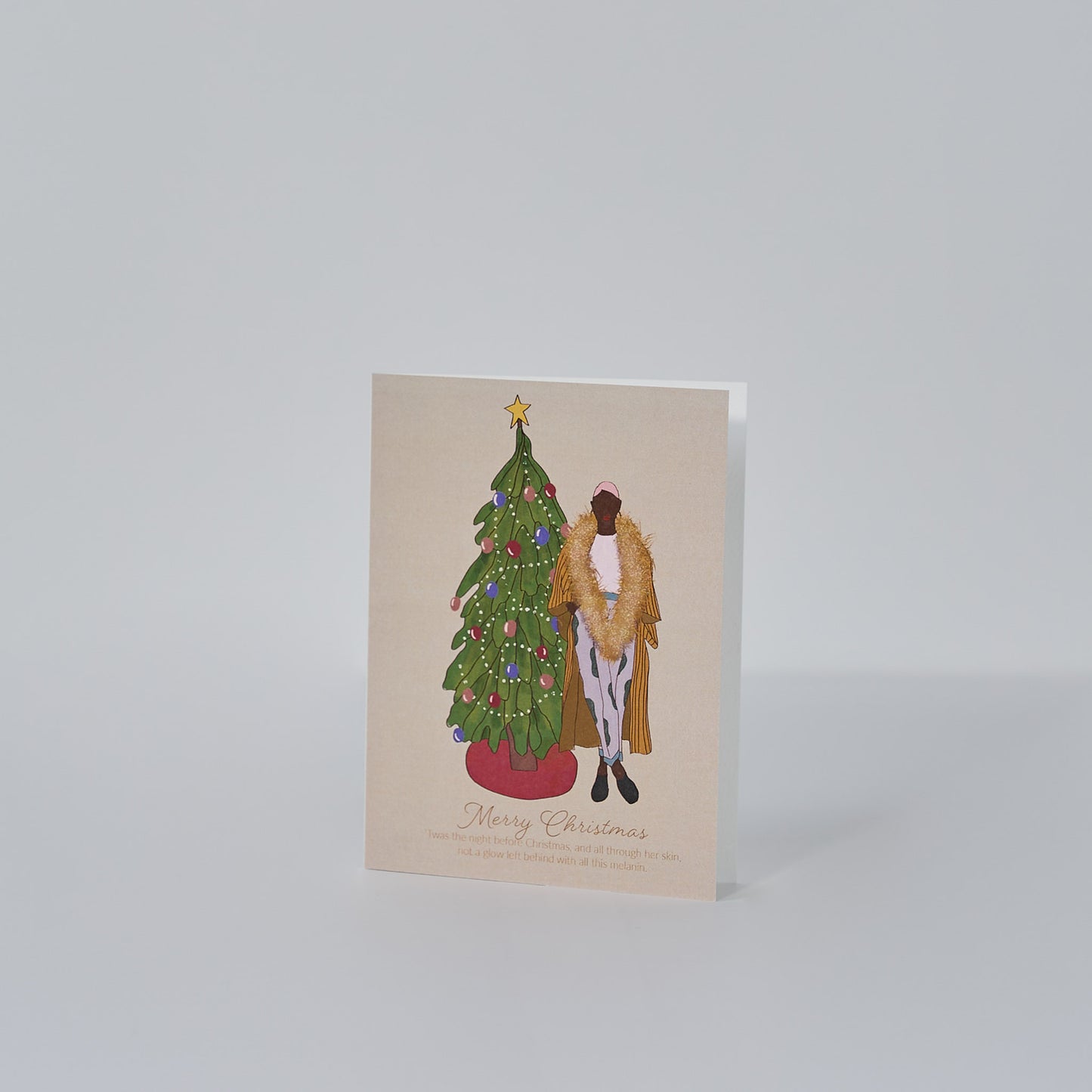 tan greeting card that reads merry christmas with illustration of christmas tree, colorful bulbs, red tree skirt, and a black woman standing in holiday attire with a large gold garland around her neck