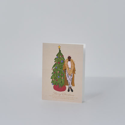 tan greeting card that reads merry christmas with illustration of christmas tree, colorful bulbs, red tree skirt, and a black woman standing in holiday attire with a large gold garland around her neck
