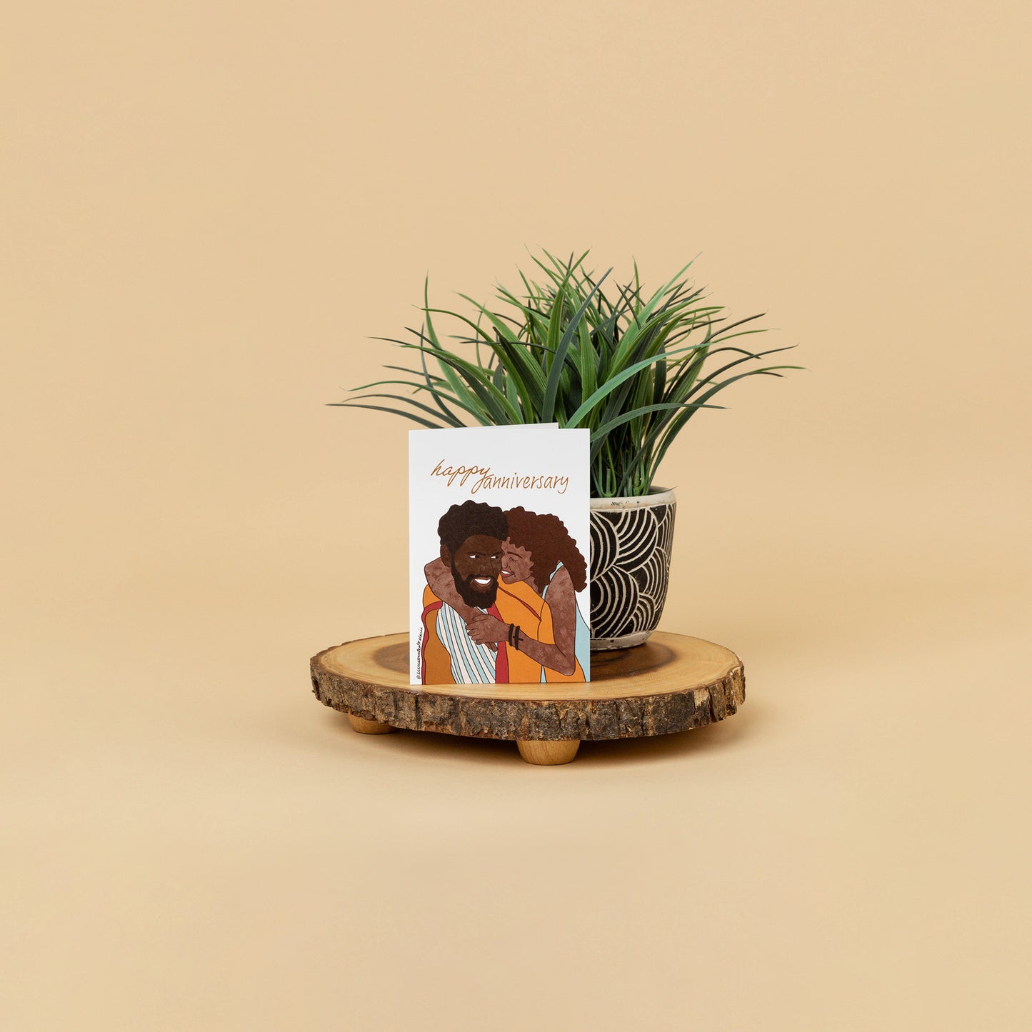 styled greeting card scene of black couple embracing each other with full smiles while card stands on wood decor against a house plant