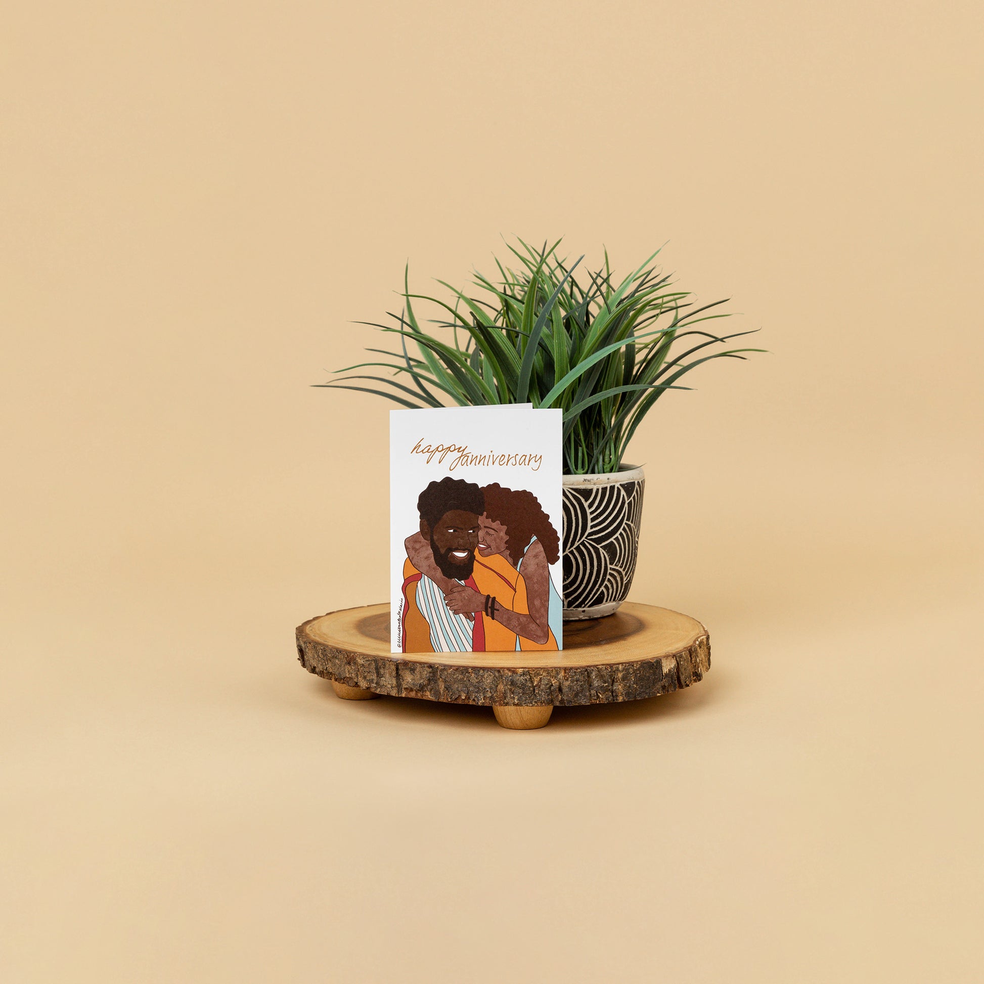 styled greeting card scene of black couple embracing each other with full smiles while card stands on wood decor against a house plant